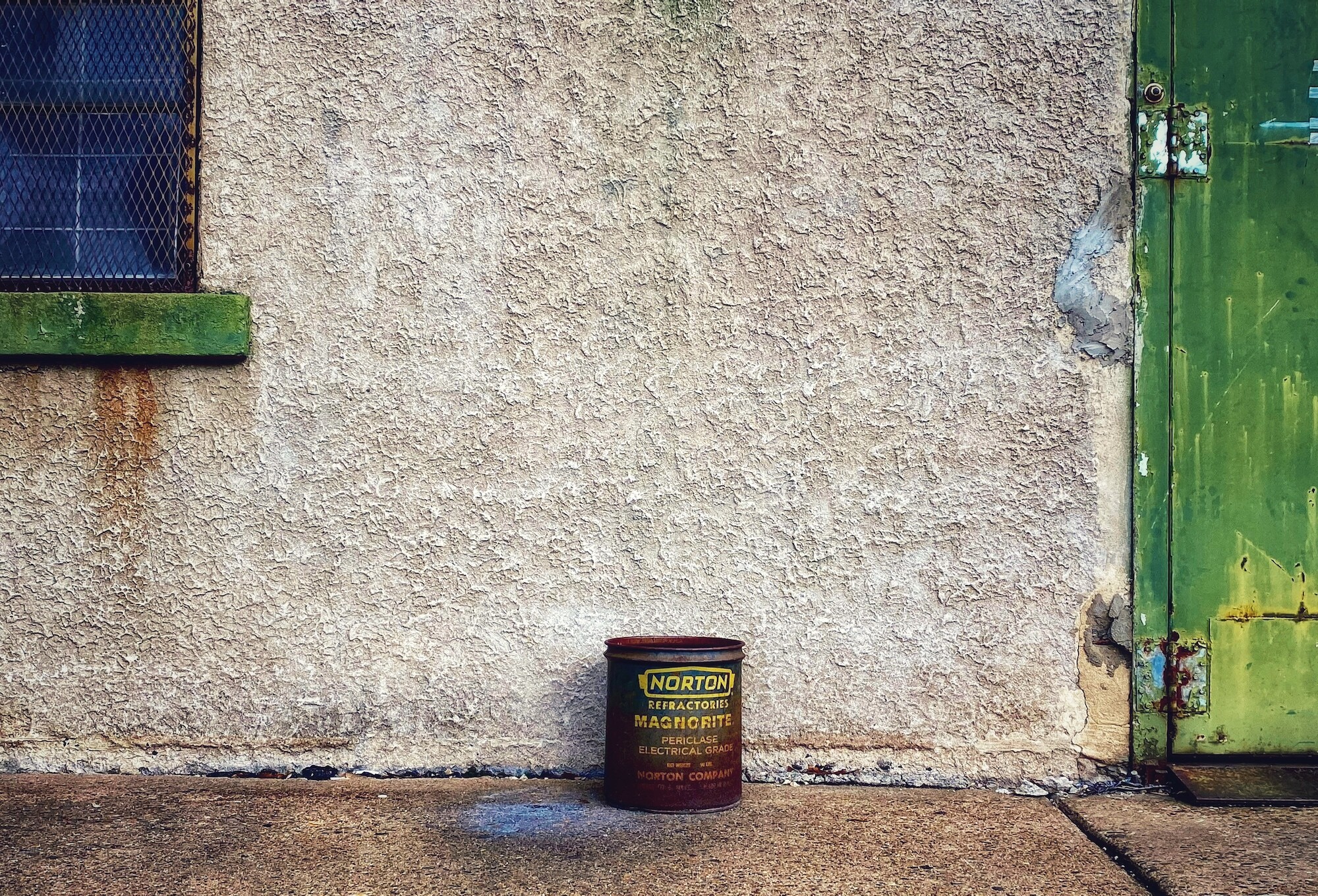 A large can in the middle of the image. The outside reads, "Norton Refracteries Magnorite Petriclase Electrical Grade, Norton Company." To the right of the can is part of a green door. To the left of the can is part of a window with a green sill and wiring across the front.