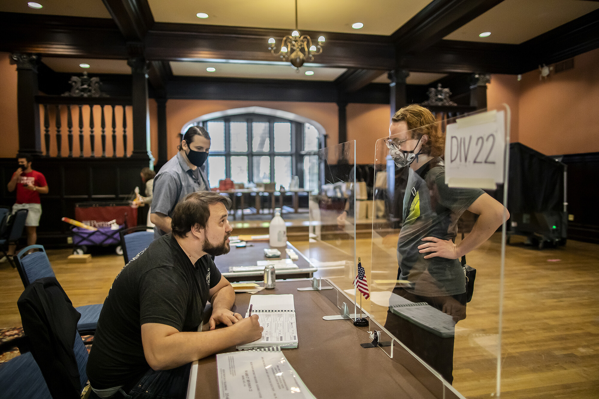 Poll worker sits at a desk in Houston Hall behind a glass partition talking to a masked voter.