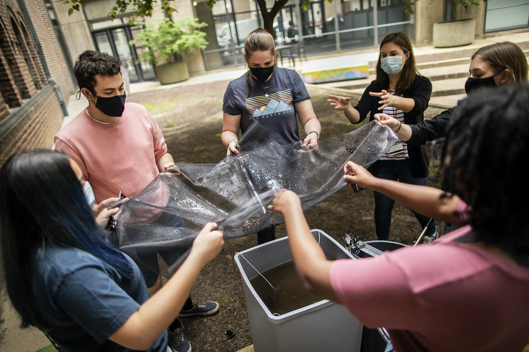 A group of five people standing outside holding a mesh net over a water basin. A sixth person is nearby gesturing with hands near the netting. All are wearing masks.