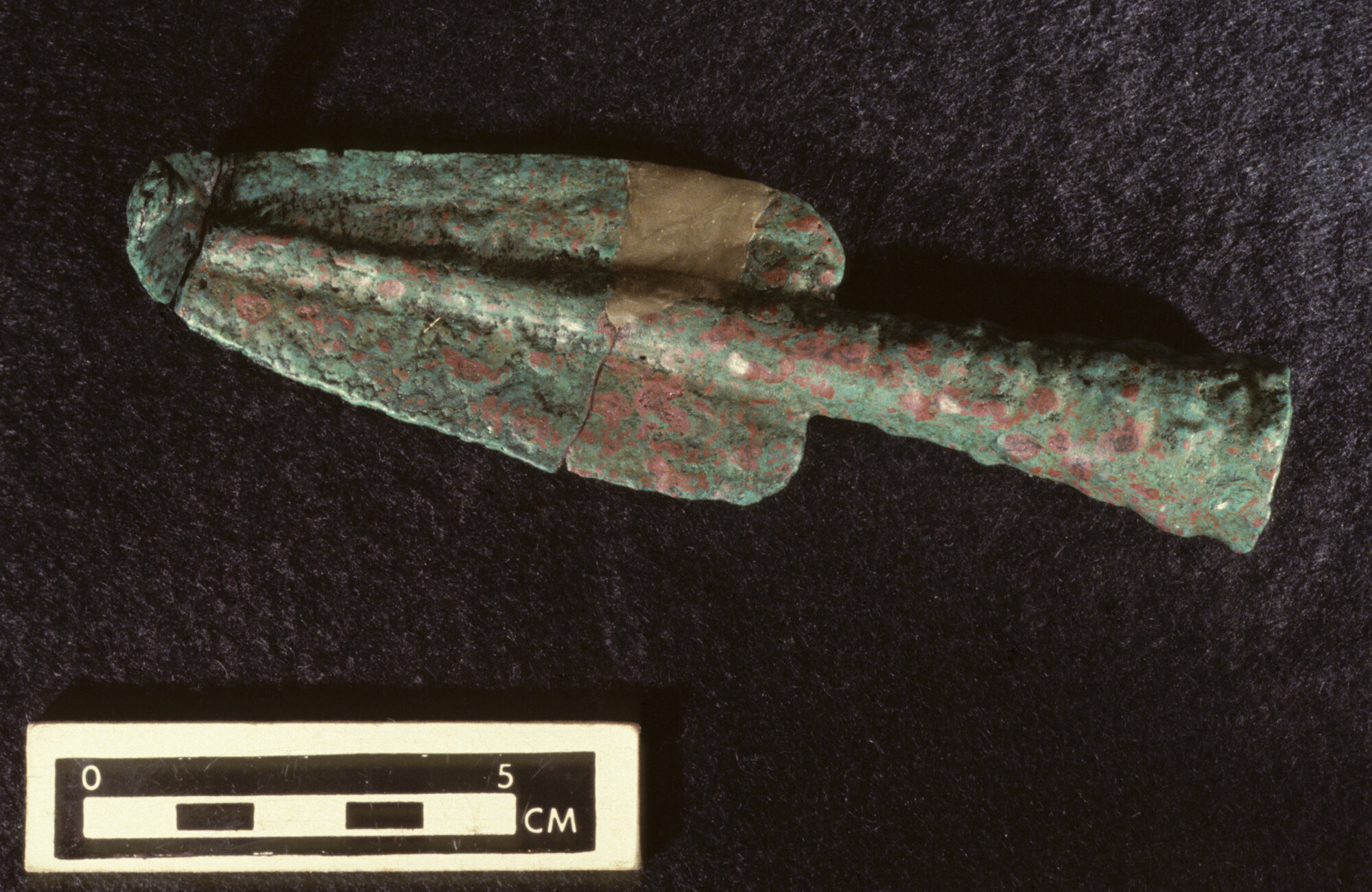 A photo of a metal artifact in the shape of a spear on a black background. In the foreground is a scale that runs from 0 to 5 cm.
