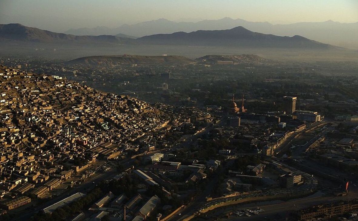 A city in the desert is seen from above, with brown structures dotting the valley and mountain ranges in the distance, some covered in haze.