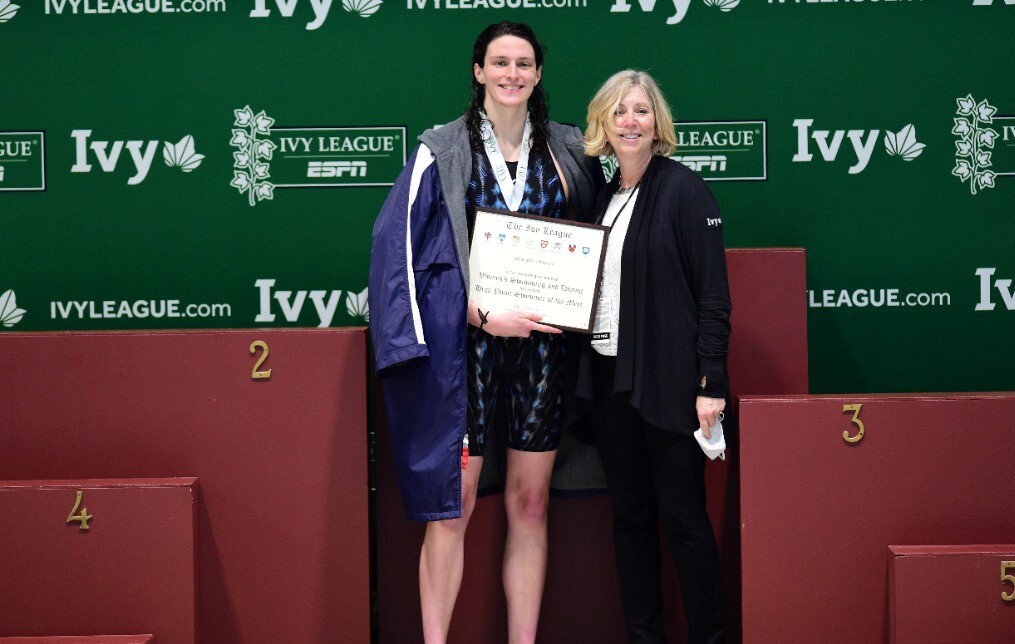 Lia Thomas holds her championship plaque while standing on the podium alongside an Ivy official.