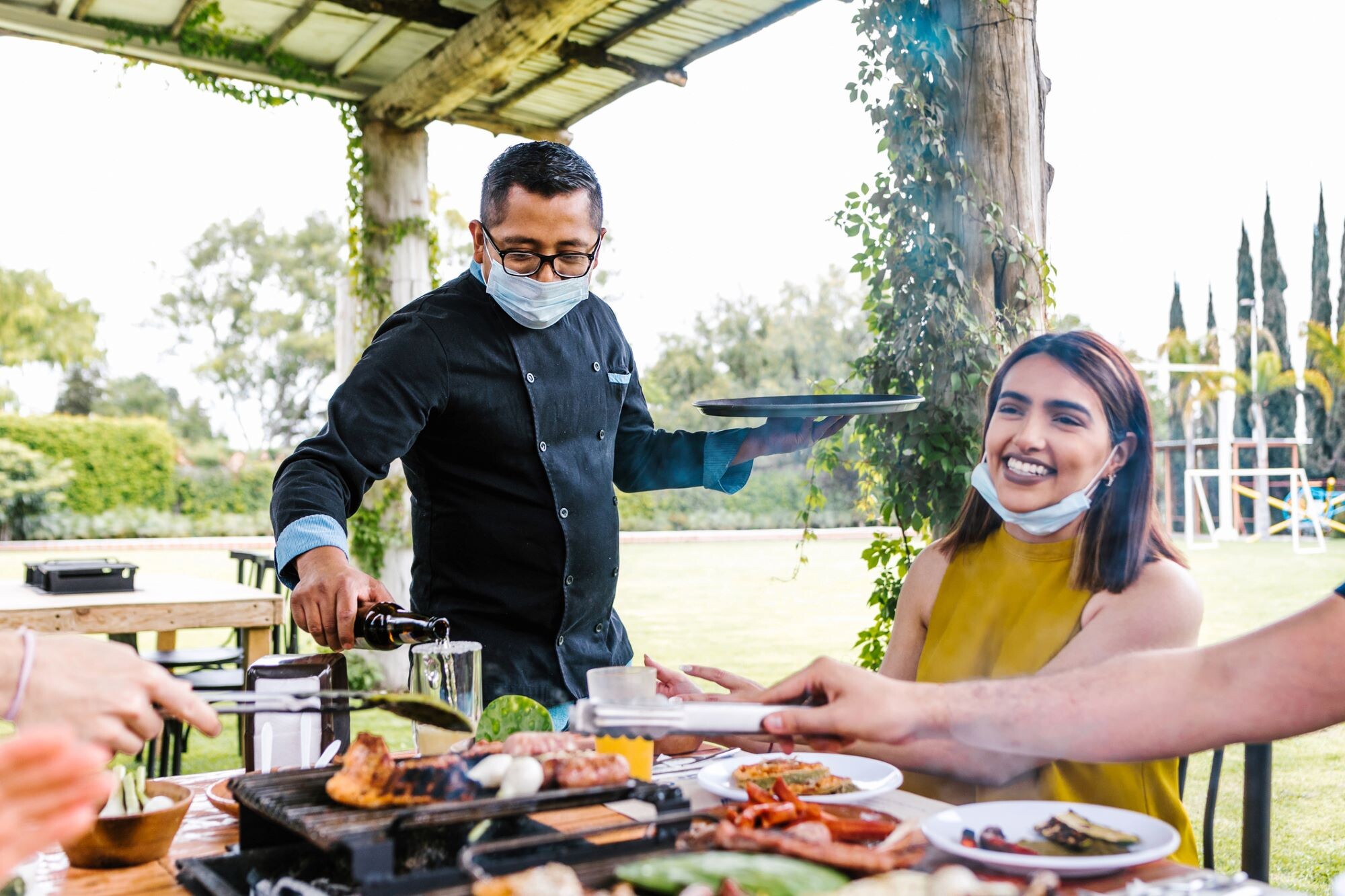 On a sunny day on an outdoor patio, a man in a surgical mask pours a smiling woman a drink.