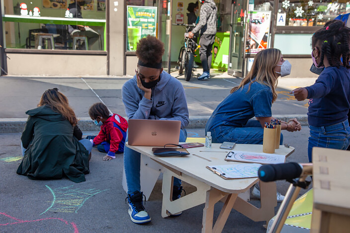Students in a parking lot of a school, some playing, one on a laptop, during the pandemic.