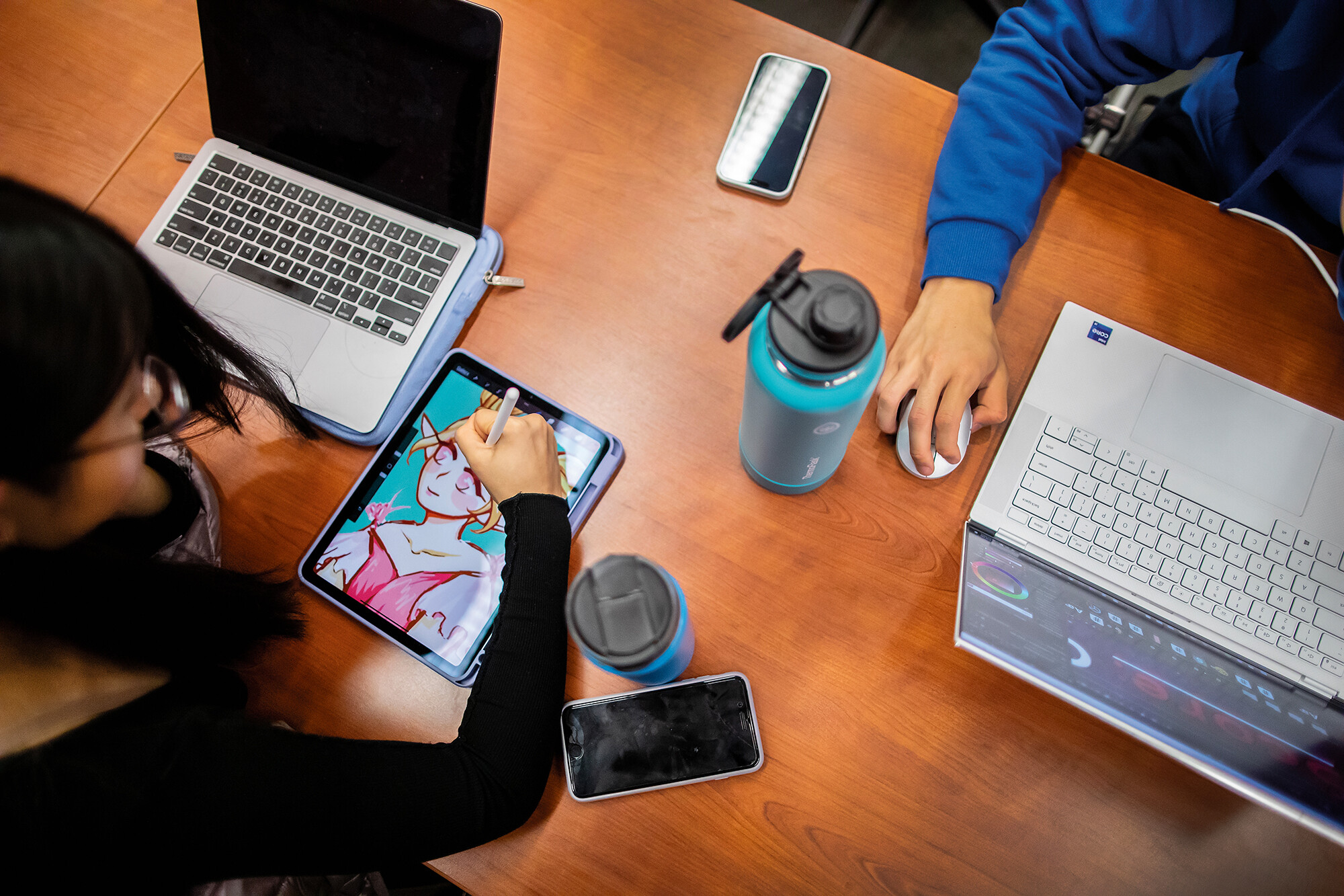 Two students at a table with laptops, one is drawing a video game image on an iPad.