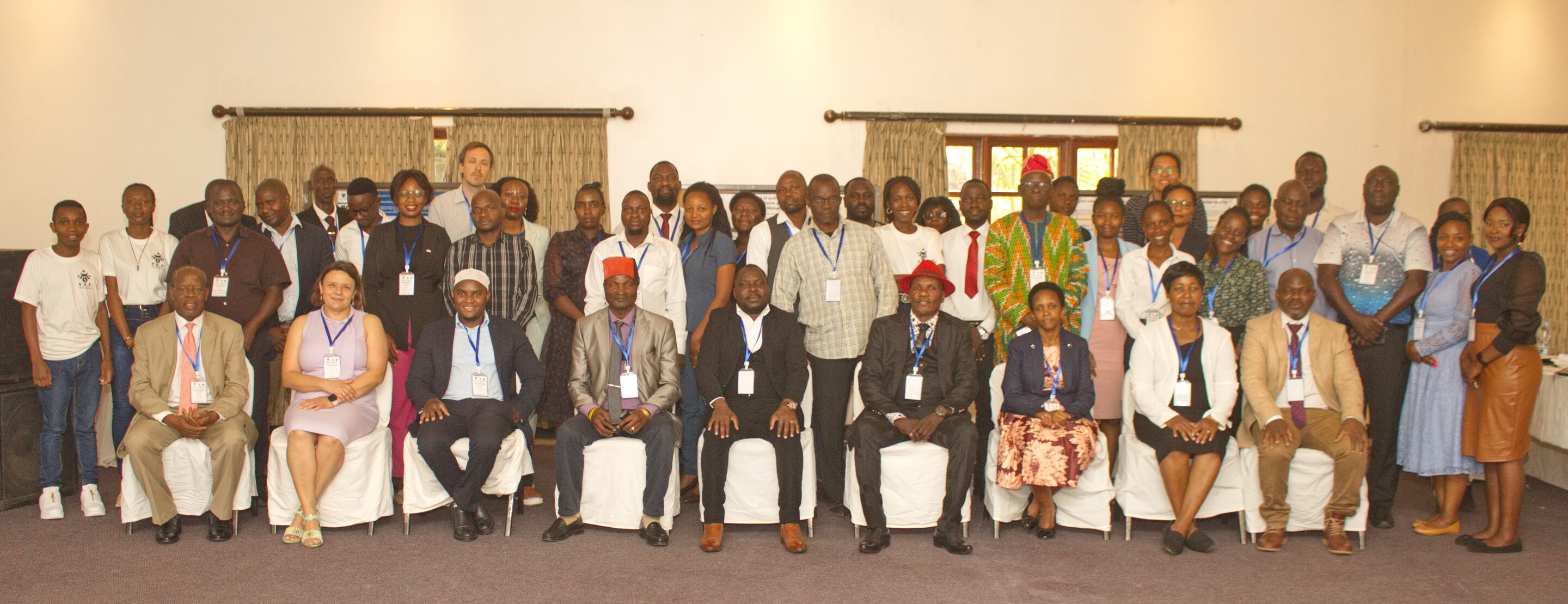 A large group of people, one row sitting, another two rows standing, in a conference room at a hotel.
