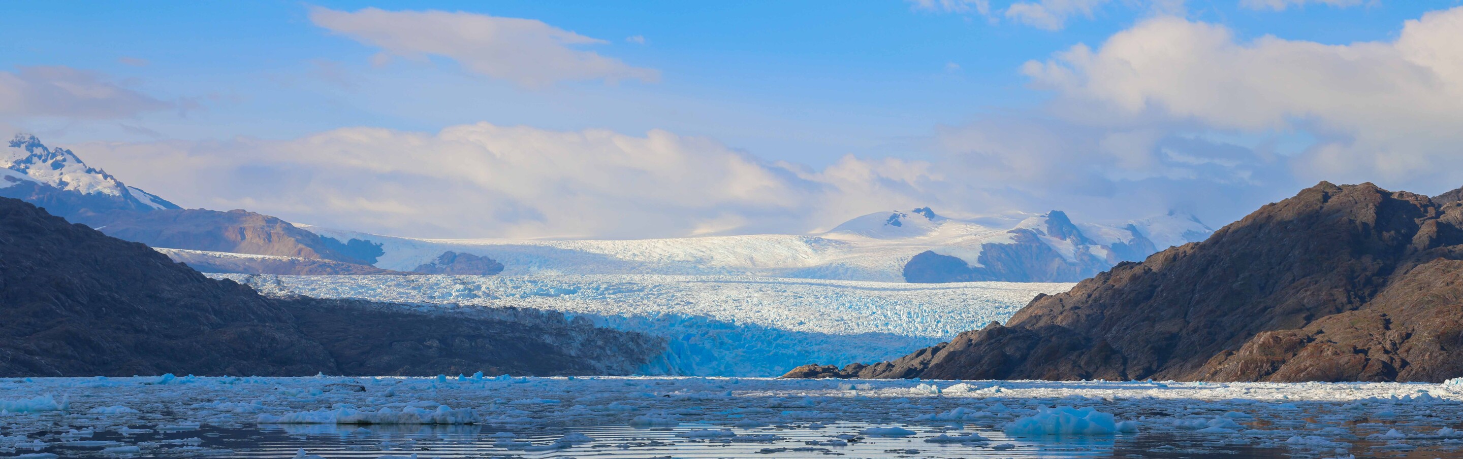 Large glacier ends in a watery pool with snowy mountains in the distance