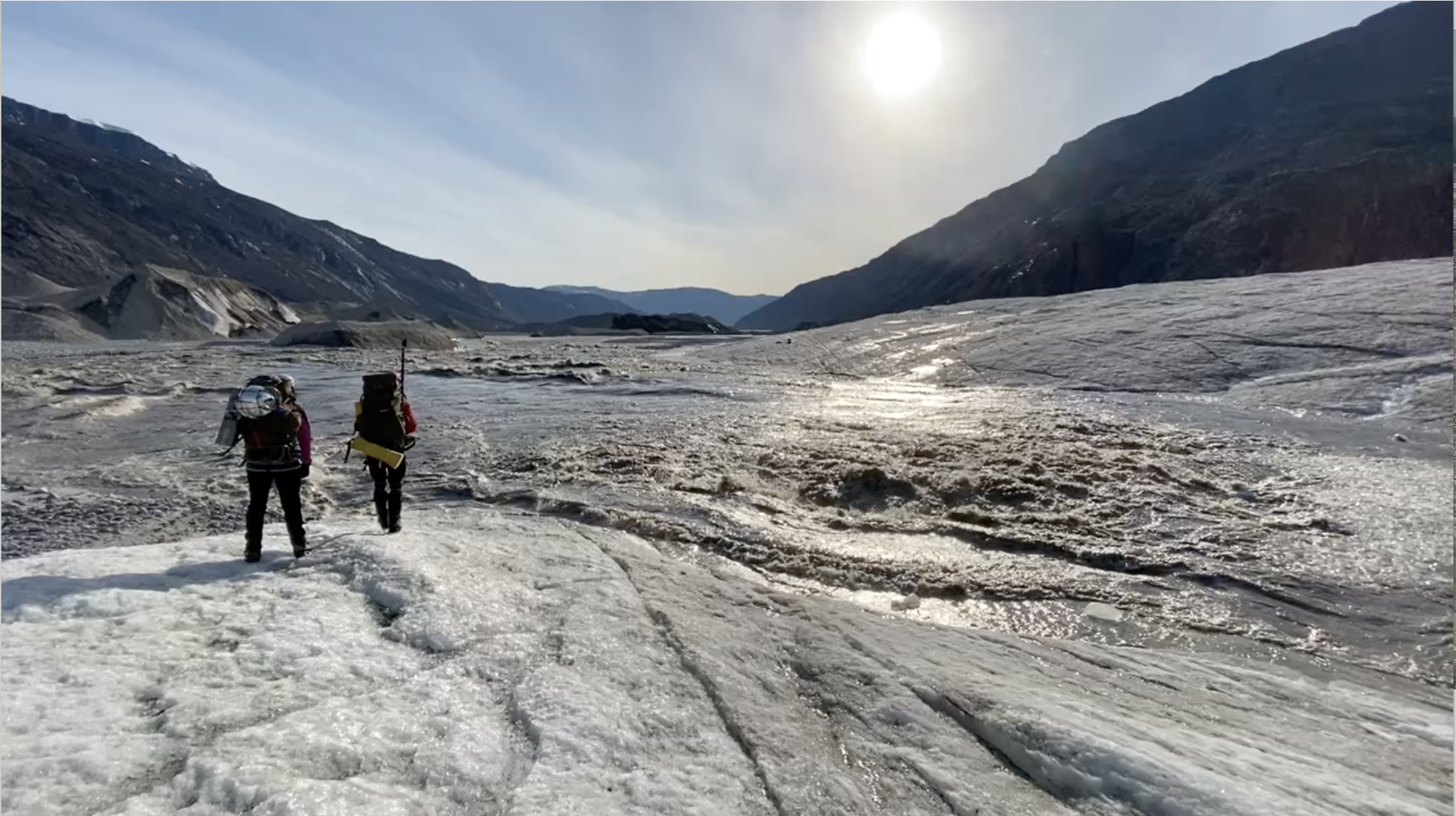 Two scientists walk on glacier ice near a river and mountains