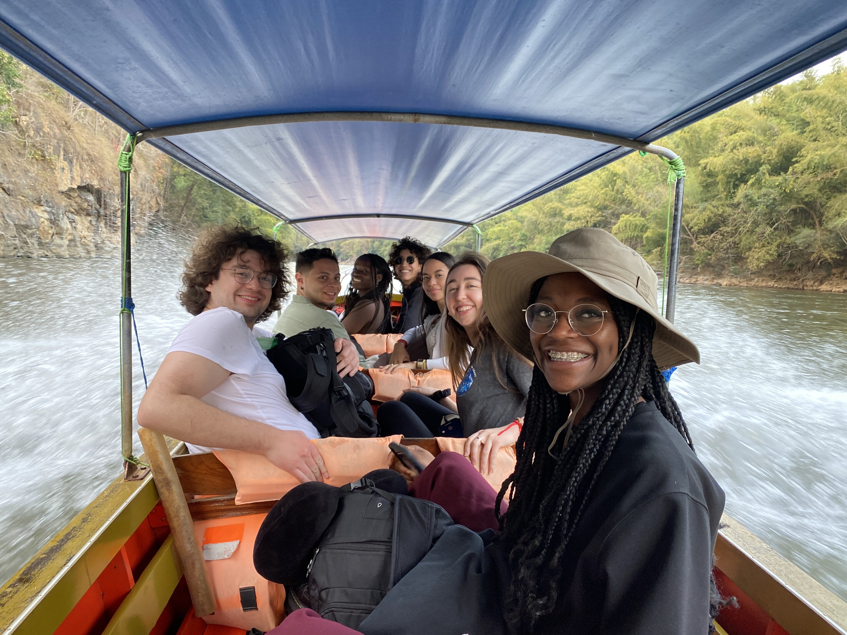 Seven students smile under the canopy of a motor-powered boat travelling on a river