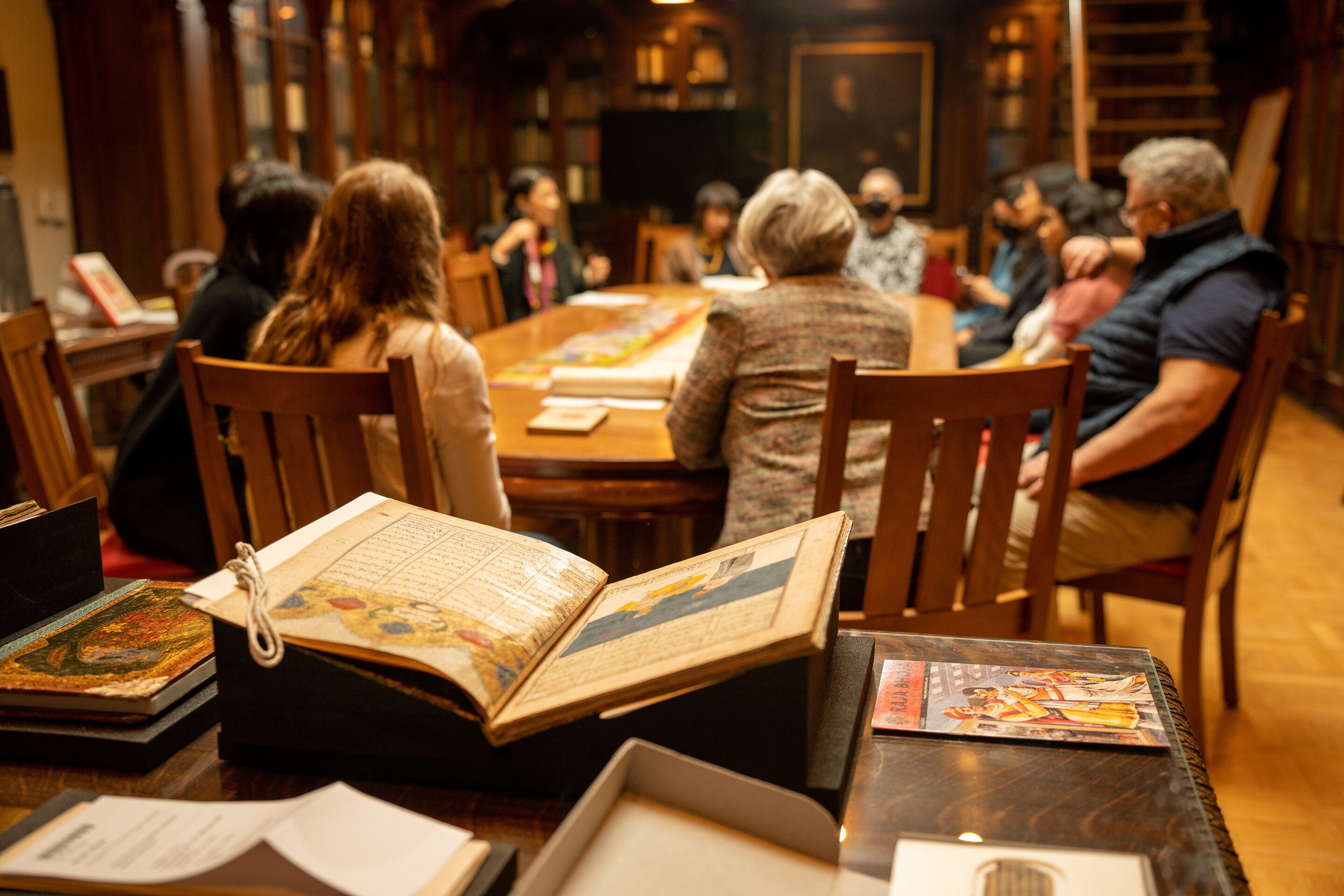 A group of people seated in wooden chairs around a large table. Books are spread out on the surface. 