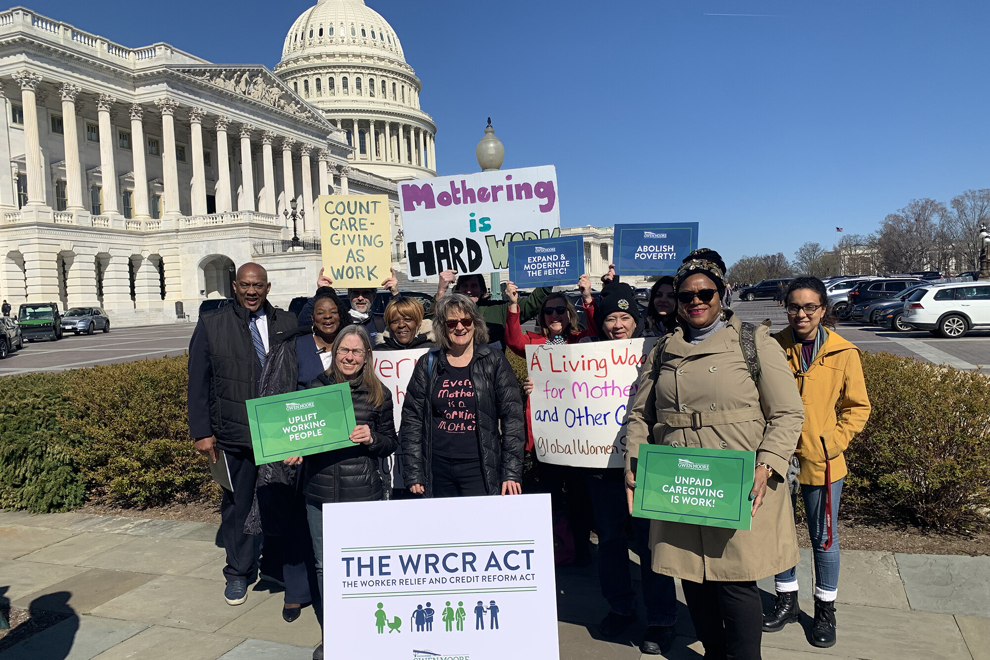 People carrying signs reading "The WRCR Act," "Mothering is HARD WORK," and "Unpaid Caregiving is work" outside of the Capitol Building in Washington, D.C.