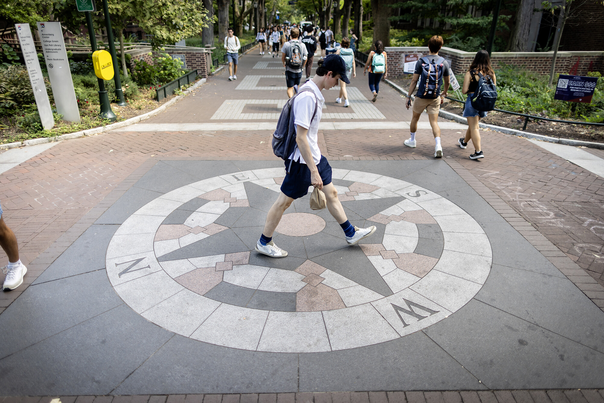 A person in shorts and a baseball hat wearing a backpack walks across a compass embedded in Locust Walk on Penn campus.