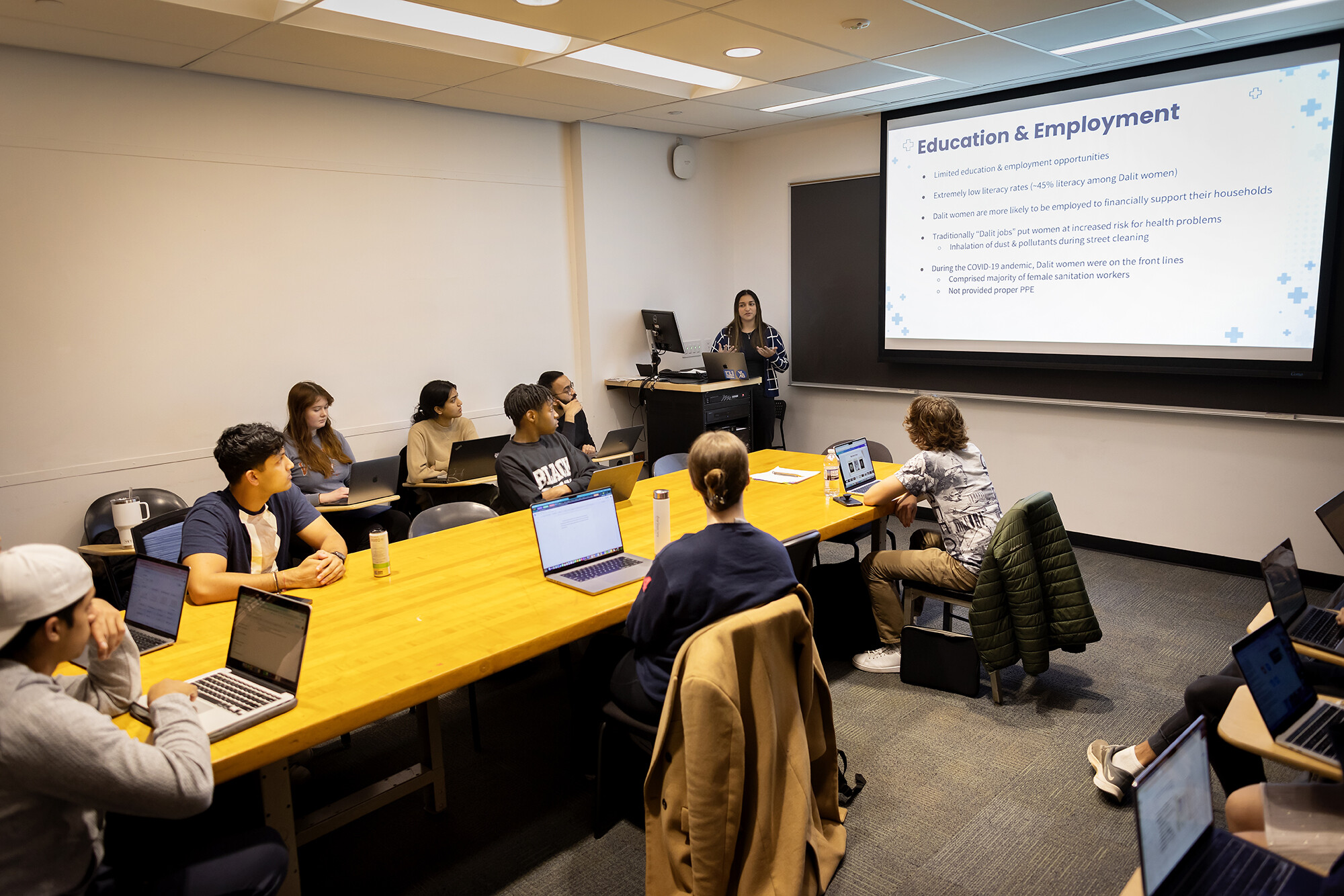 Class listens to a presentation. The presenting student is at the front of the class discussing "education & employment"