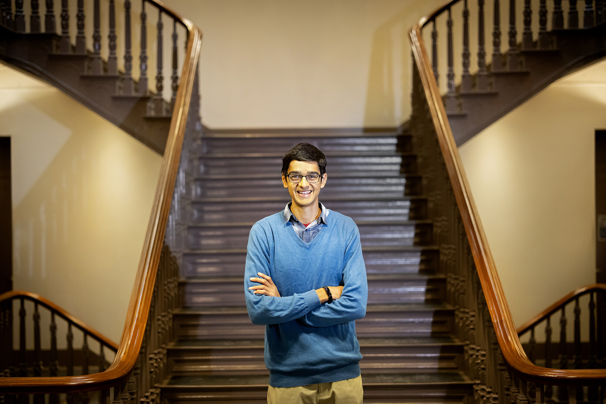 Wearing glasses and a blue sweater, Om Manghani stands with crossed arms on a staircase