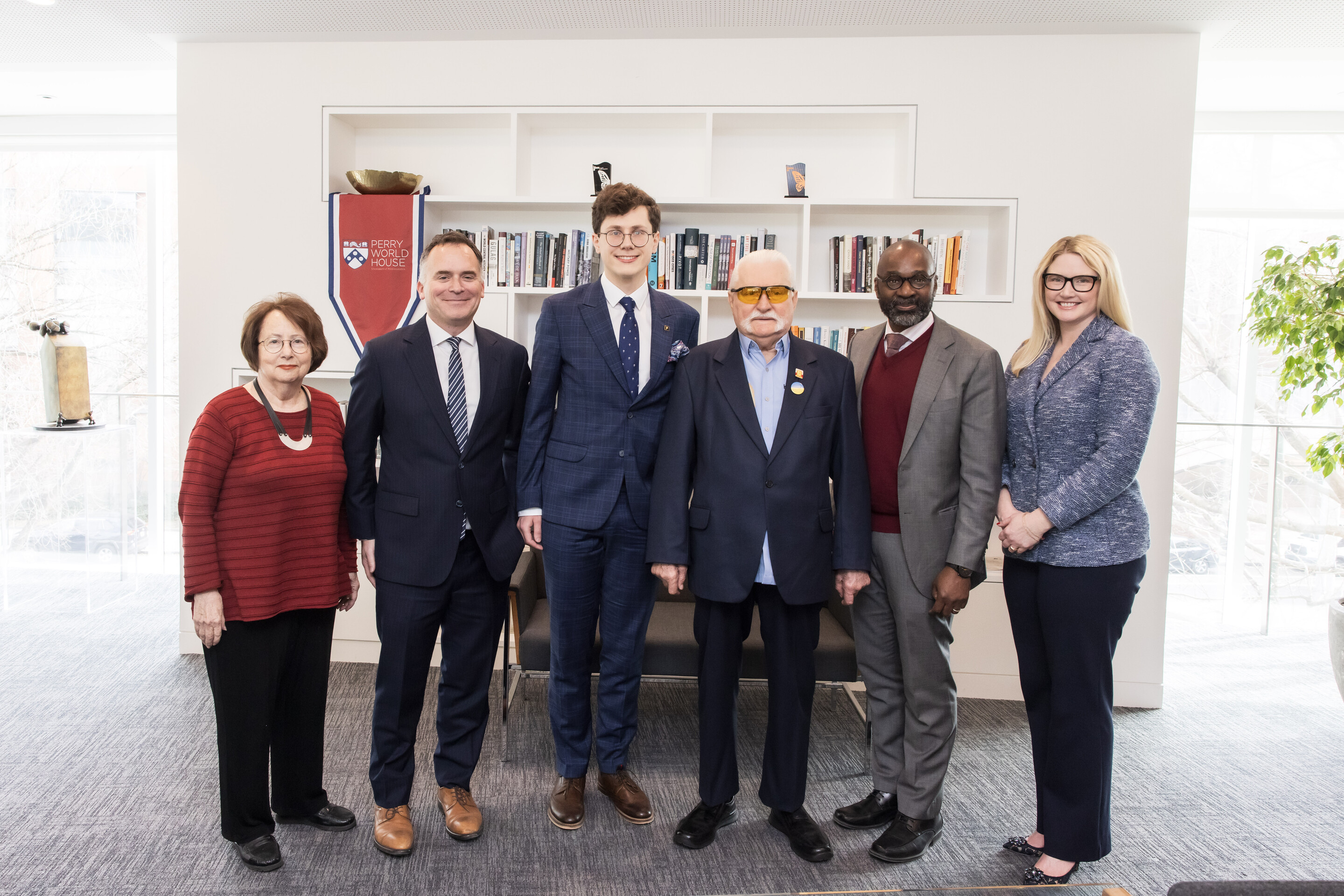 A group of six people stand in front of a bookcase and a white background with a Penn Perry World House banner.