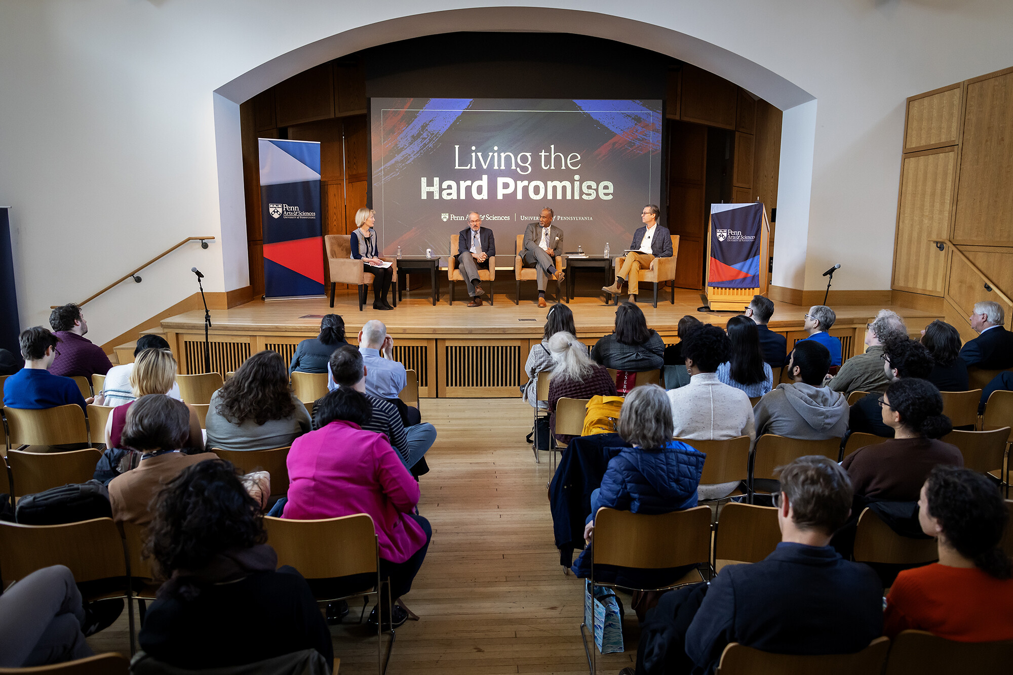 Four people sit in chairs on an auditorium stage in front of a sign reading Living the Hard Promise.