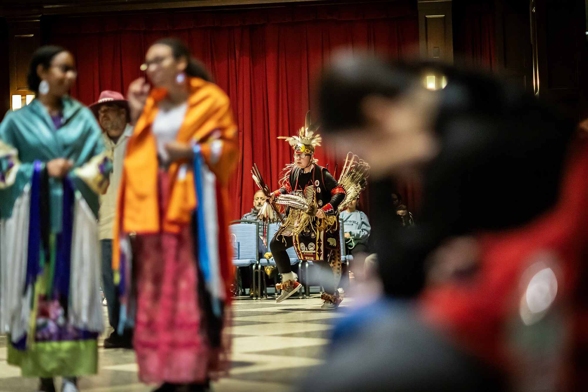 In the foreground, female dancers with shawls. In the background, a young man in warrior regalia. 