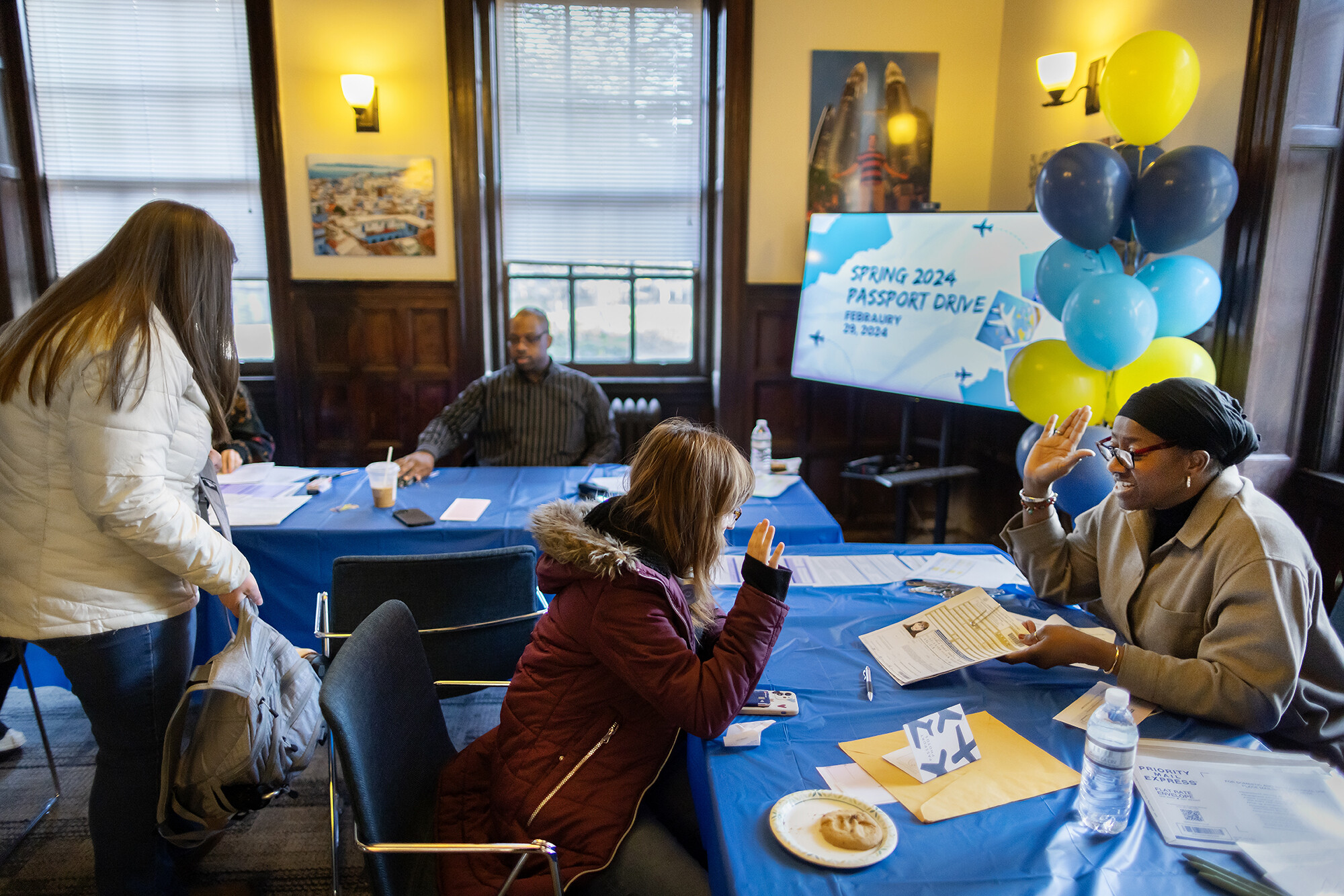 People sitting at tables covered in blue table cloths, with the two people in the foreground facing each other, each holding up their right hands as one person asks the other to take an oath for a U.S. passport.
