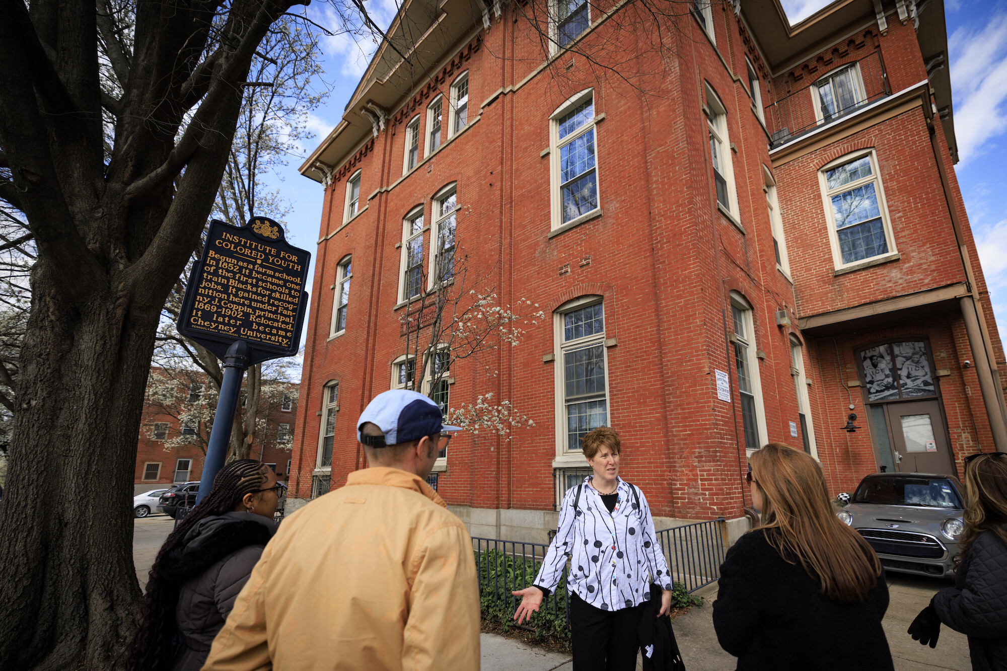 Amy Hillier addresses a small group in front of a brick three-story building. A plaque out front reads: Institute for Colored Youth