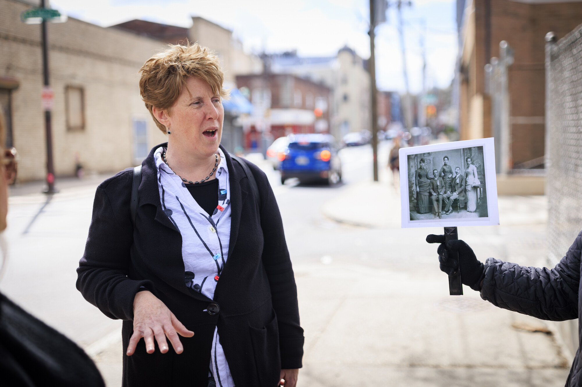 Amy Hillier speaks on a street in Philadelphia. To her right, a hand holds up a black-and-white historic photograph 