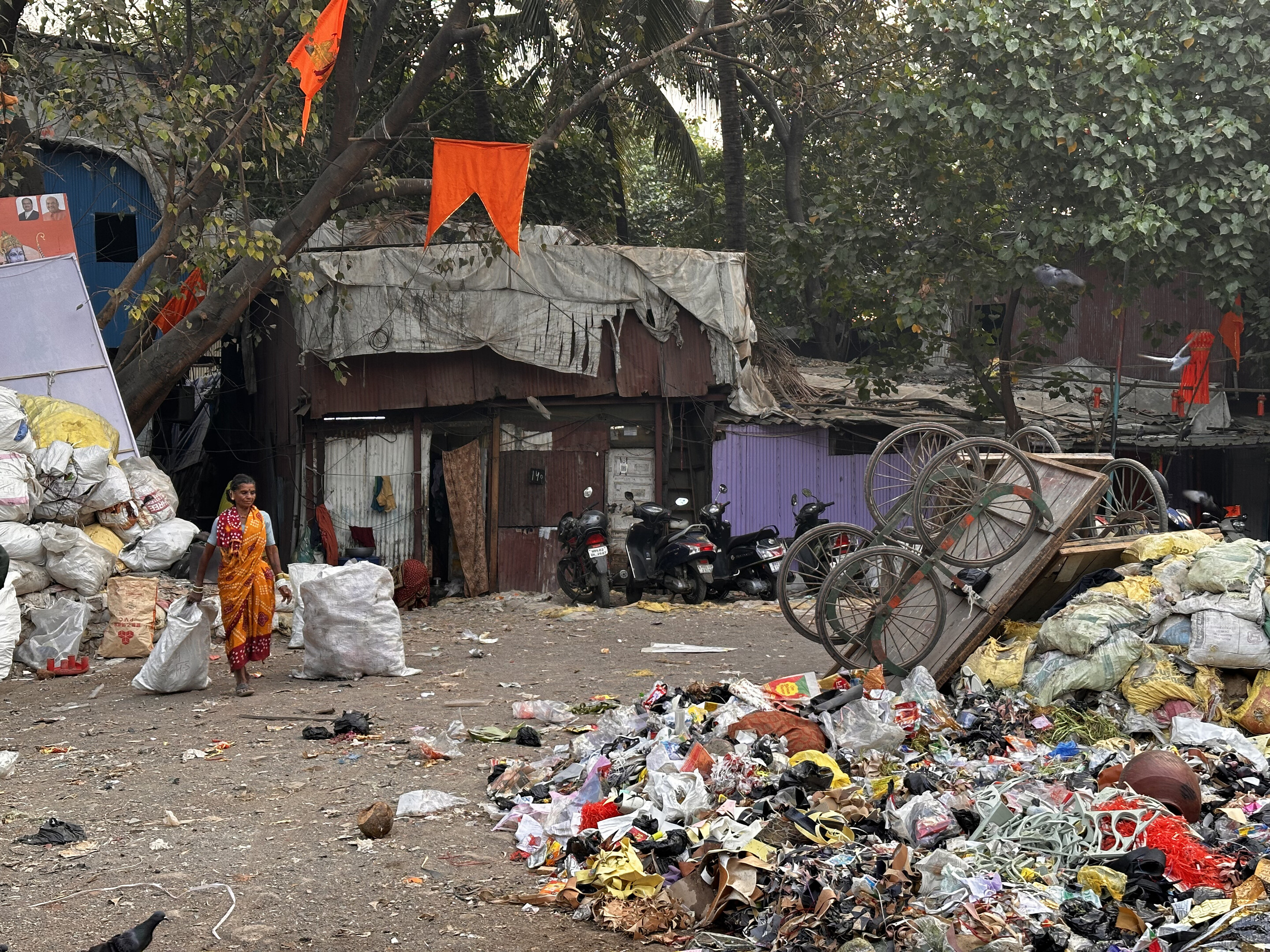A woman in an orange sari walks through an informal settlement, dragging two large plastic bags with her. 