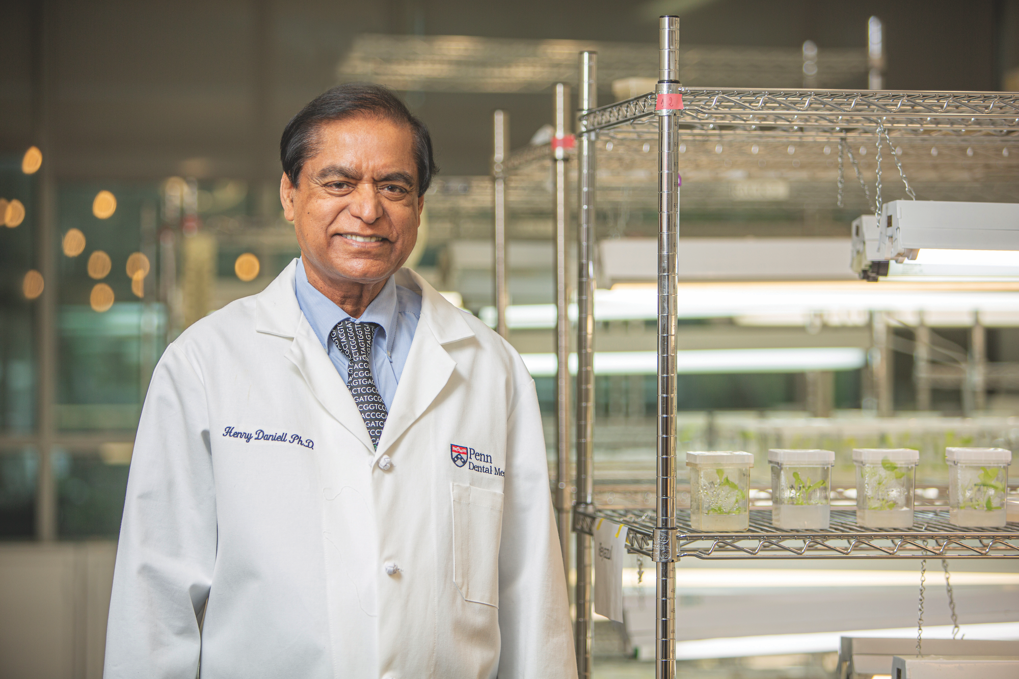Daniell stands in in lab coat in his lab next to shelves with small plants in jars.