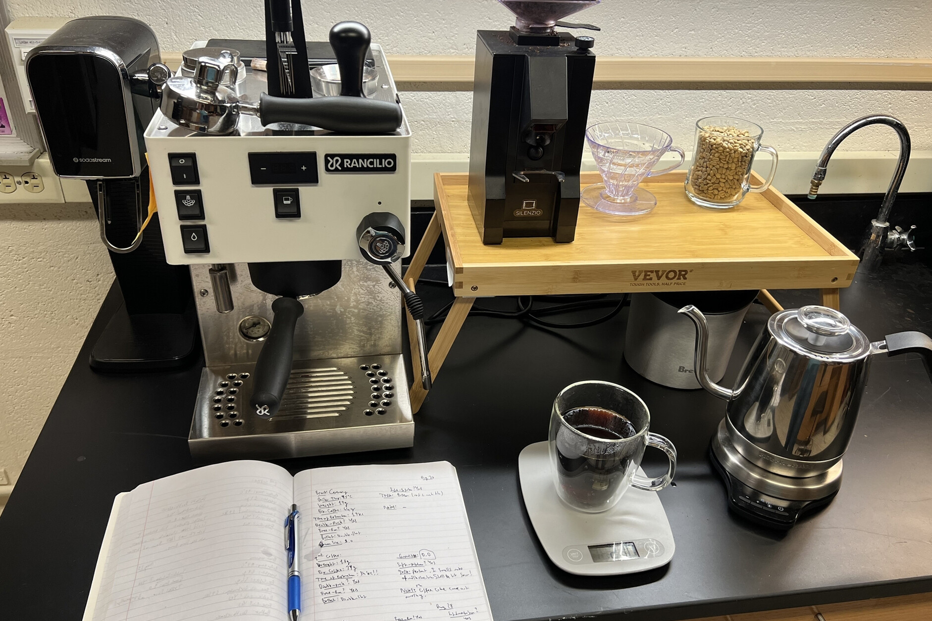 A coffee machine, kettle, scale, cup of coffee, and notebook in a lab.
