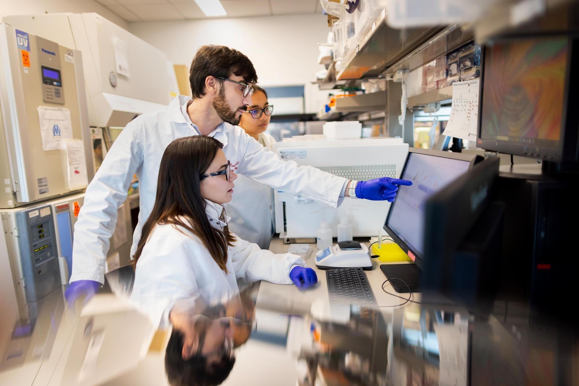 Marco Ruella in the Ruella Lab, collaborating with two lab technicians. Ruella is wearing a white lab coat and pointing at a computer screen.