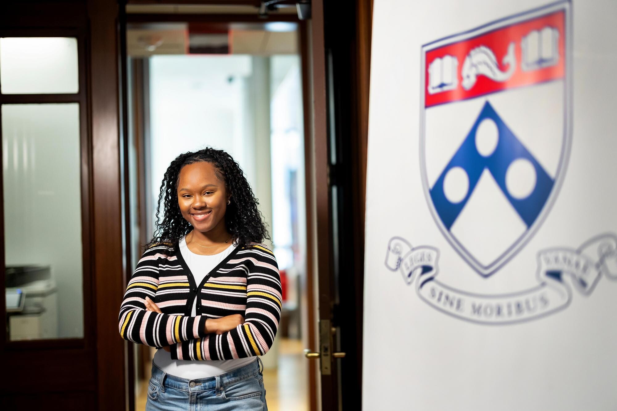 Mikhaidia Miller wearing a striped sweater and standing with arms crossed, smiling and facing forward, in the Penn First Plus office. Penn's logo is on the right.