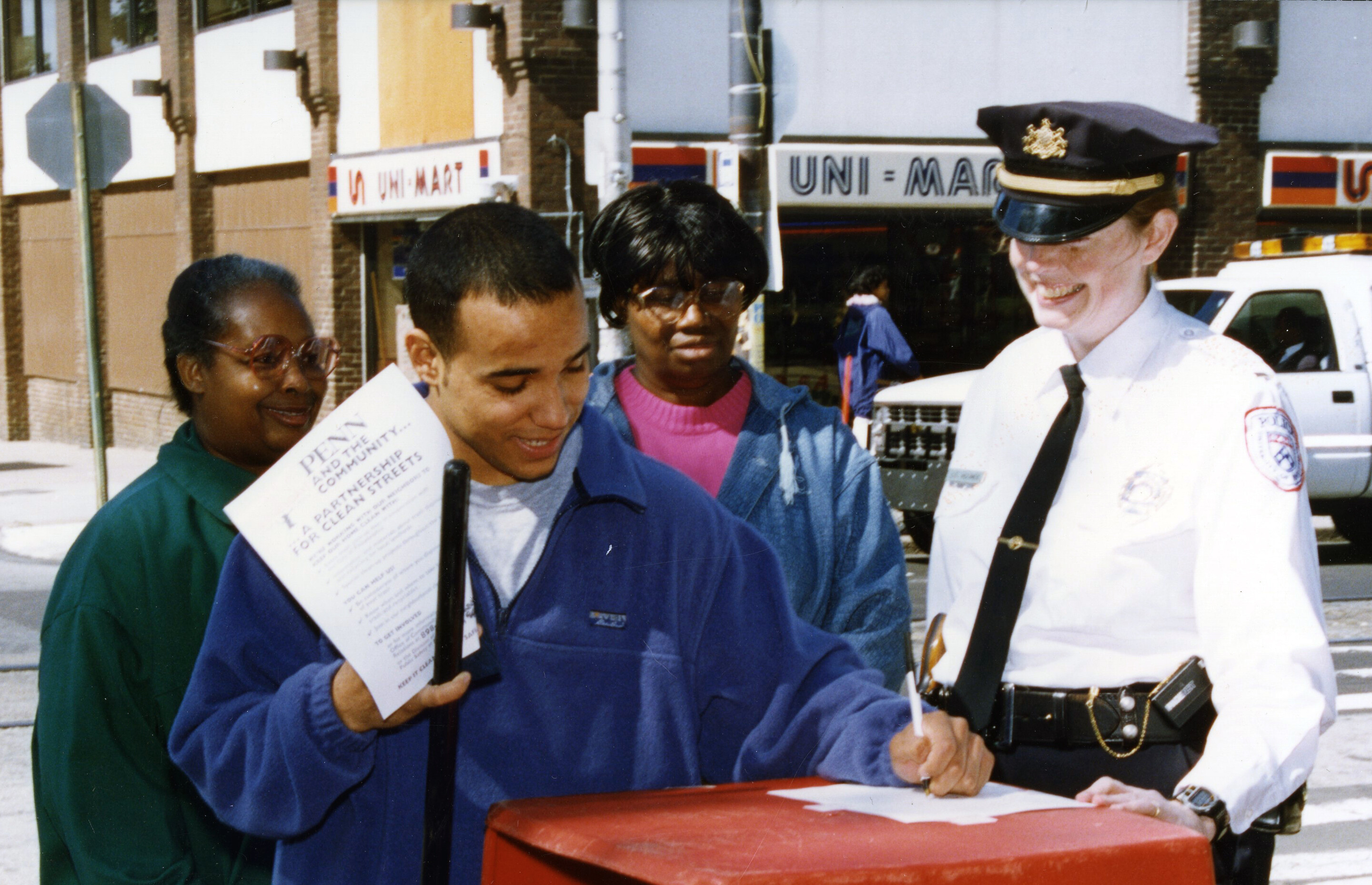 Three West Philadelphia community members interacting with a policewoman near a garbage container in an outdoor street setting. A young man is writing on a piece of paper.