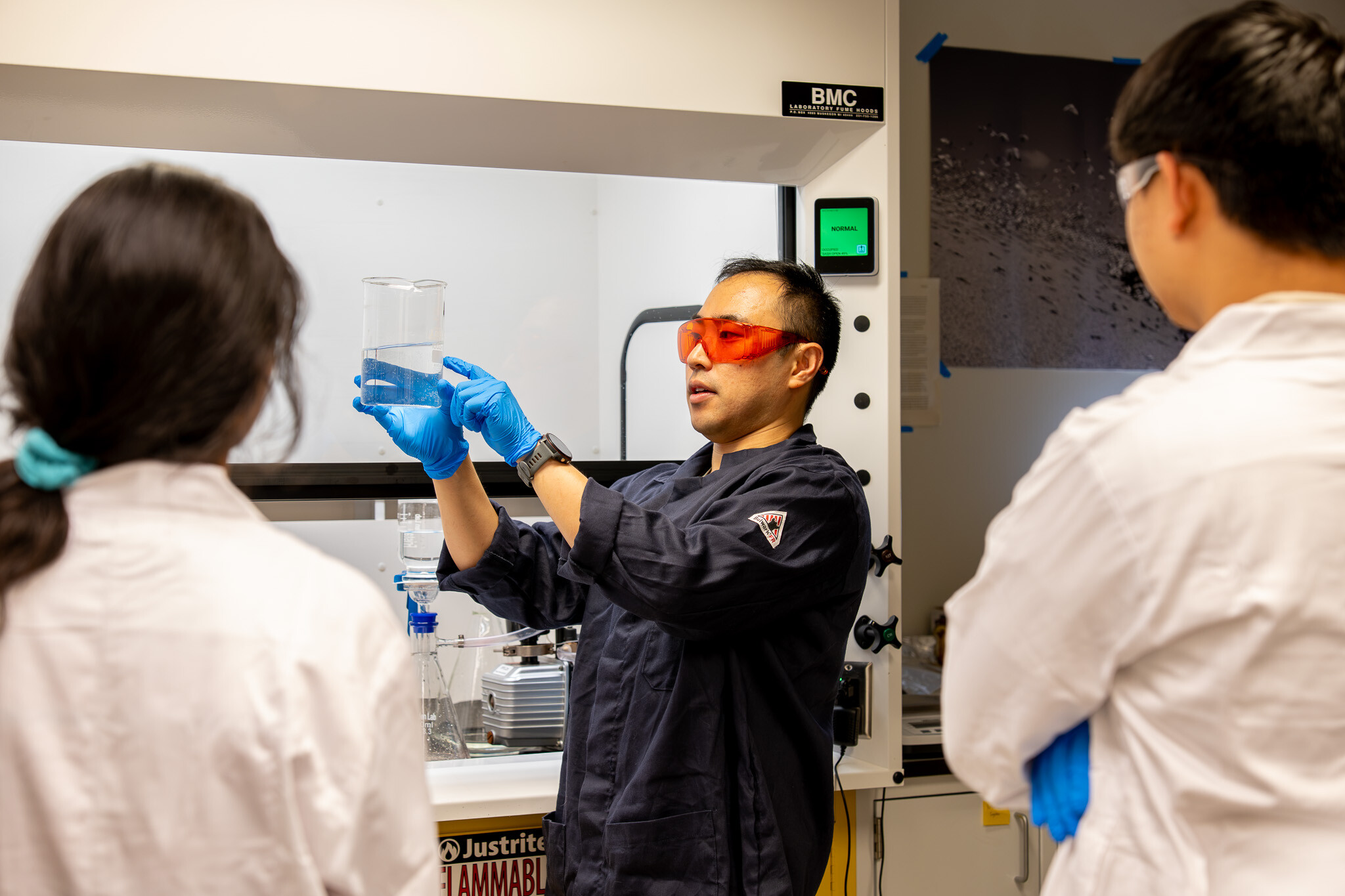 Derek Ho in the lab pointing to a beaker of water in front of two lab techs.