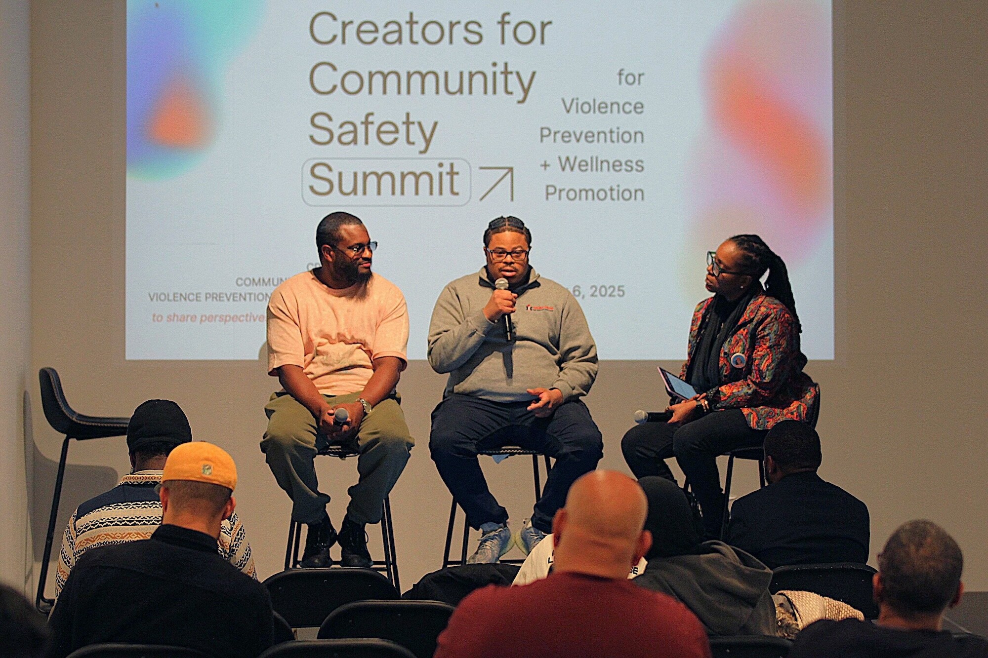 Creators for Community Safety Summit panelists Jaime Meekins (left) and Rodney Babb (center) speaking with facilitator Tamara Russell (right) on stage. The slideshow in the background reads: "Creators for Community Safety Summit" and "for violence prevention + wellness promotion"