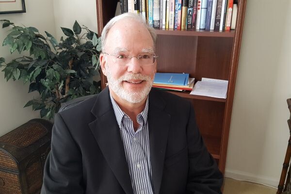 A person sitting in an office wearing a navy blazer and a striped button-down shirt. Behind are a bookcase with a shelf of books, a plant, and a wicker chest.