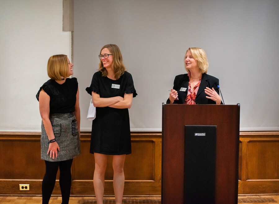 one woman at a podium gesturing with her hands while talking and turning to other women who are reacting with smiles