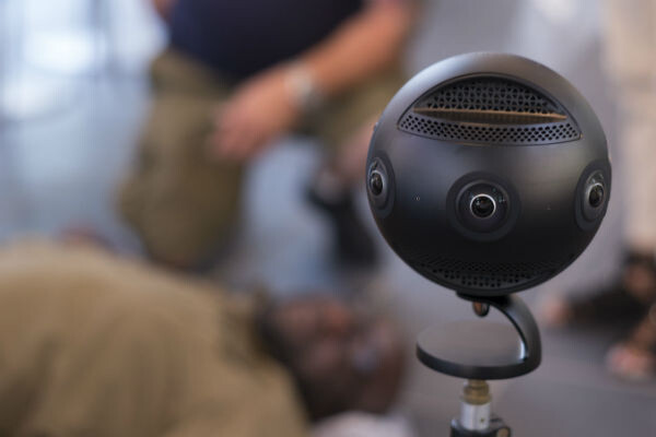 A black, six-lens camera in the foreground, with actors blurred in the background.