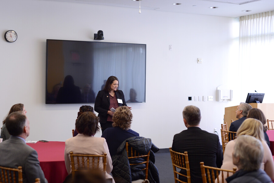 A speaker stands in front of a screen giving remarks to an audience of several people seated at round tables