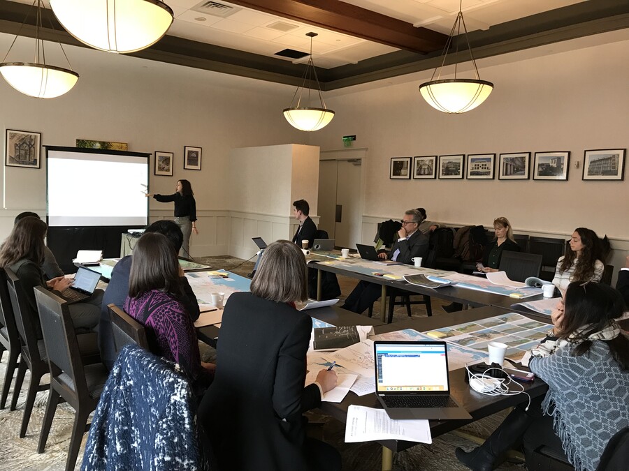 Meeting attendees around a horseshoe shaped table pay attention to a presentation at the front of the room.