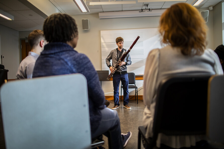 A man plays a bassoon, with two women and a man watching. The observers' backs are to the viewer. 