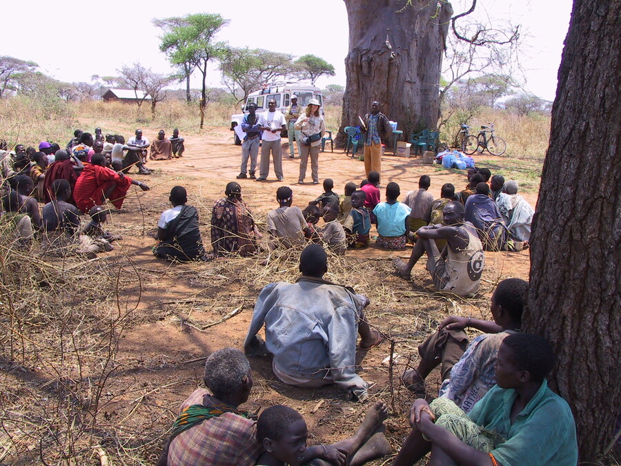 A large group of people sit on the ground outside, roughly in a circle around a group of presenters in the African landscape.