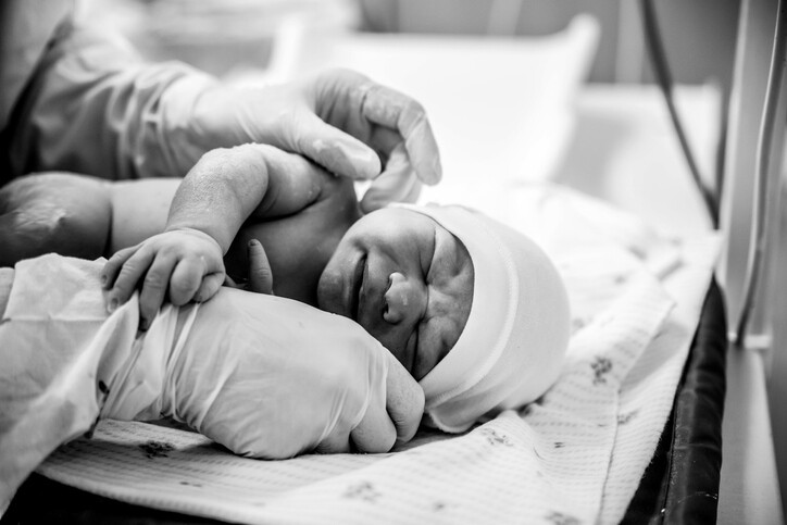  A premature baby wearing a hat is laying down in a medical setting with medical personnel wearing latex gloves touching the baby’s head