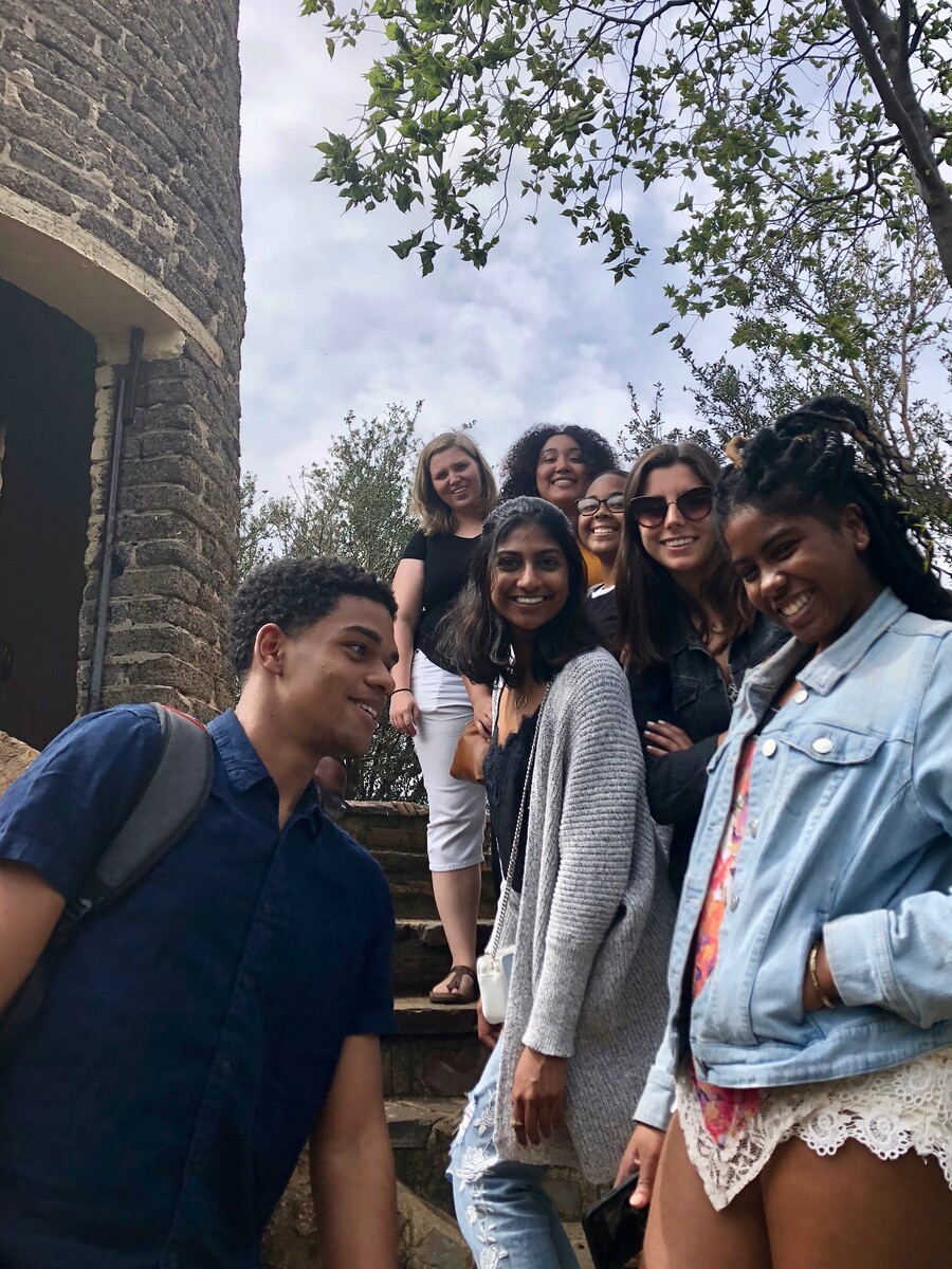 Seven students looking at camera standing on stairs next to a brick tower. 