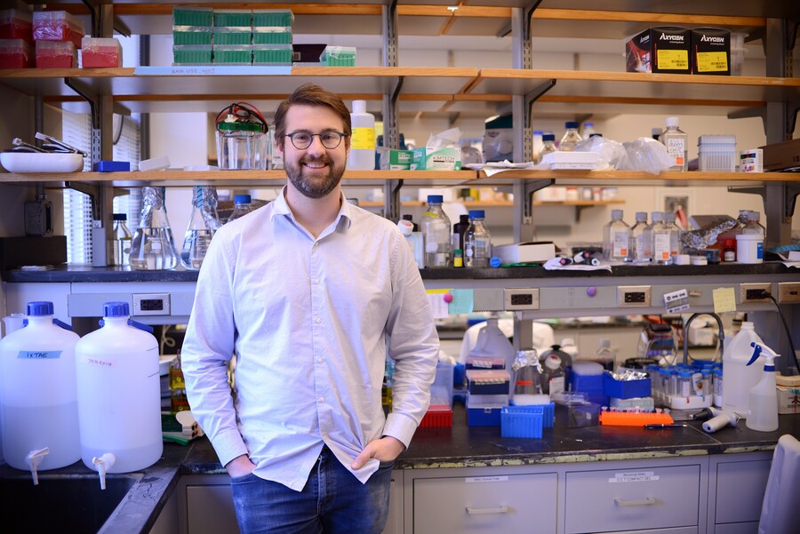 A man stands in a lab with various equipment around him: jugs of fluid, pipette tips, beakers, and more.