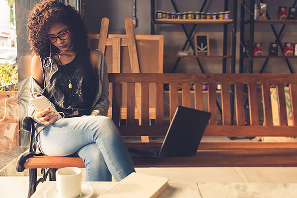 Person on a bench in a café looking at smartphone seated next to an open laptop.