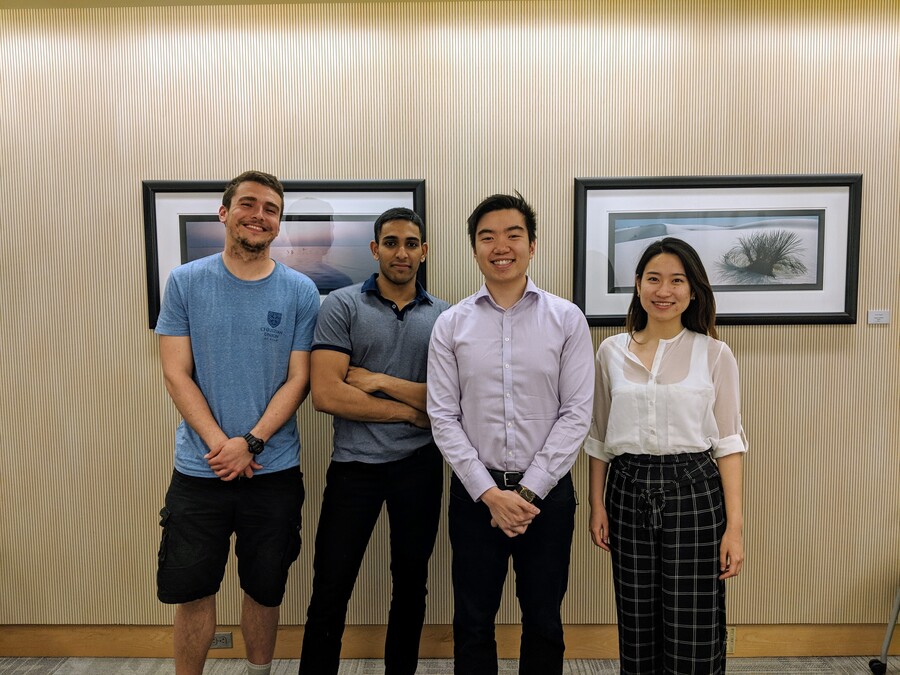 Four people standing n a row, in front of a yellowish wall with two framed images.