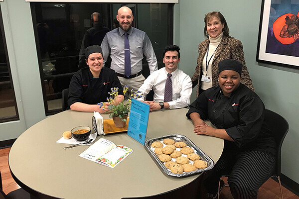 Five people seated at a table smiling, a tray of cookies in front of person wearing chef clothing.