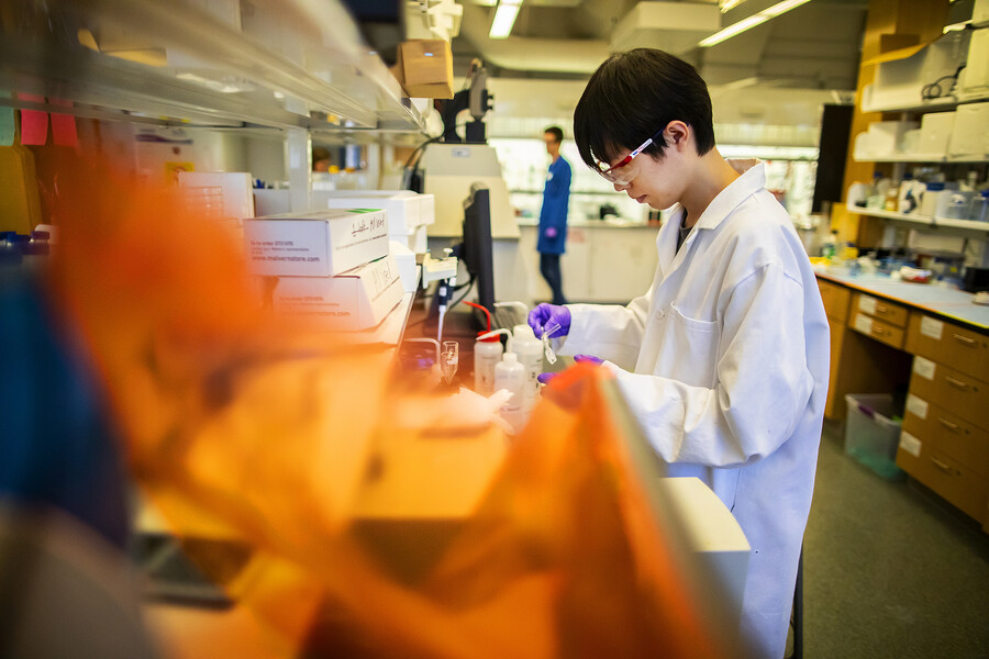 a person holding a pipette in their hand in the center of a laboratory, another person stands in the hoods blurry in the background
