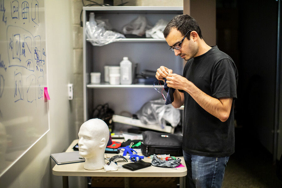 A person stands at a table in a lab covered with wires and a computer case and a styrofoam replica of a head. Across from the person is a whiteboard with drawings in blue.