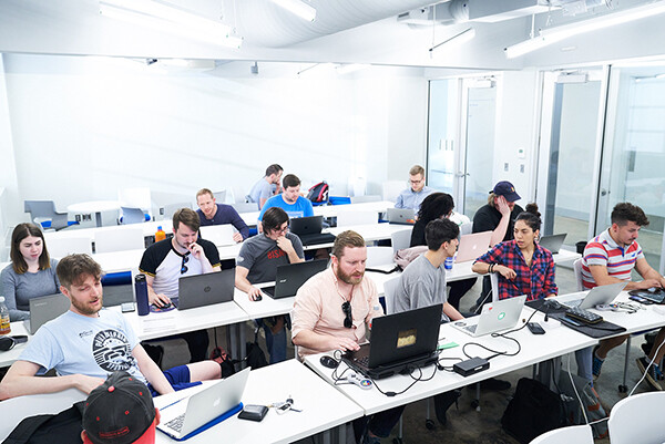A class of adults sitting at long tables working on laptops