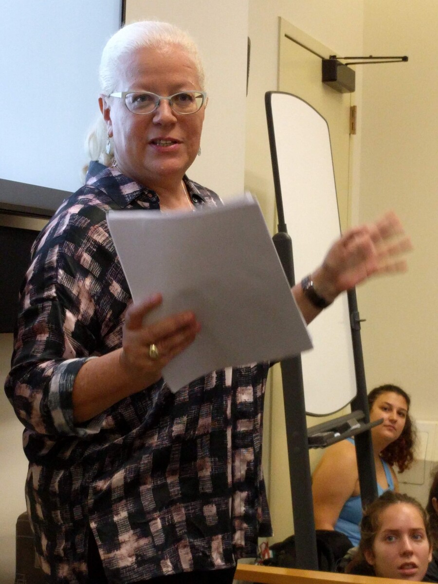 A person standing in front of a screen, holding a paper and gesturing while talking.