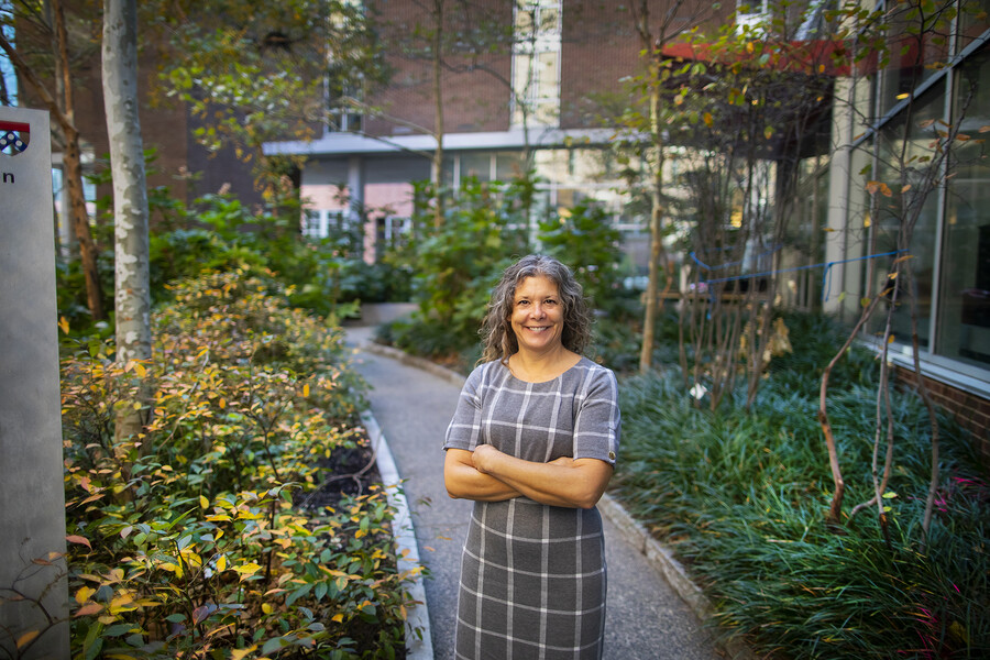 A person wearing a grey and white shortsleeve dress stands on a gravel path surrounded by shrubbery on either side. A building is visible on the right side of the image.